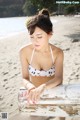 A woman in a white bikini sitting on a bench on the beach.